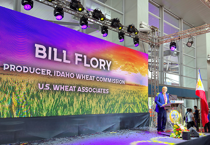 Image shows a large display and Bill Flory standing at a podium addressing a Bakery Fair in Manila, Philippines, and illustrates Flory's participation in USW activities for this Wheat Industry News post.