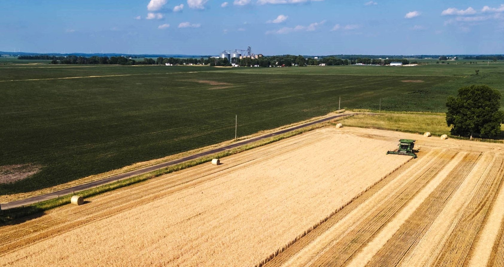 Real Ag Stock photo shows an aerial view of a combine harvesting soft red winter wheat with green fields in the background and a grain elevator on the horizon.