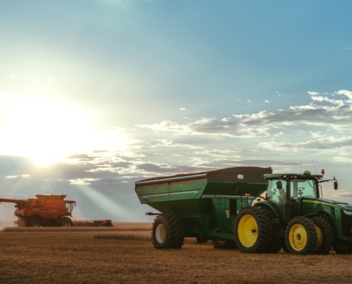 Image shows a harvester cutting wheat with a large tractor and wagon in the foreground late in a day with the sun in the background behind a cloud on the horizon.