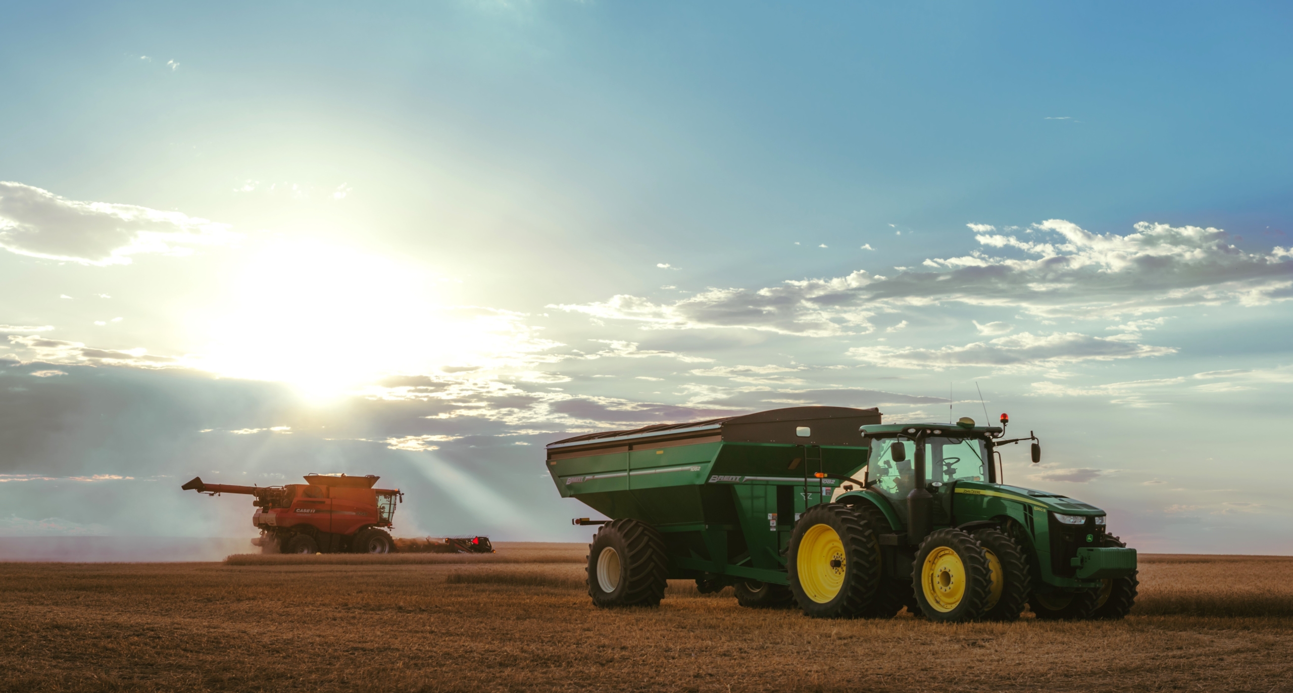 Image shows a harvester cutting wheat with a large tractor and wagon in the foreground late in a day with the sun in the background behind a cloud on the horizon.